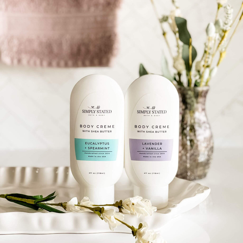 Two bottles of Simply Stated Bath & Body Aromatherapy Body Creme in Eucalyptus + Spearmint and Lavender + Vanilla, displayed on a white tray with floral accents. A soft, neutral background enhances the calming aesthetic.