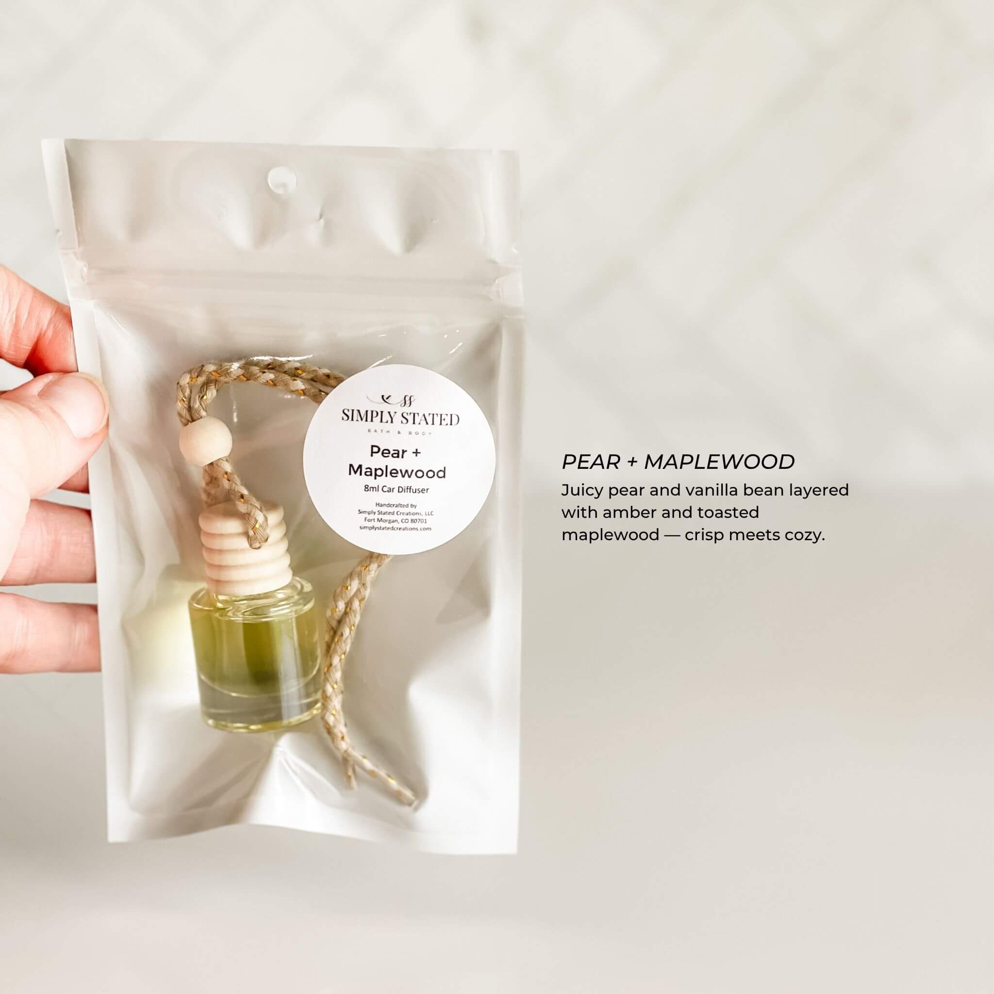 Hand holding a car air freshener with 'Pear + Maplewood' description on a plain background