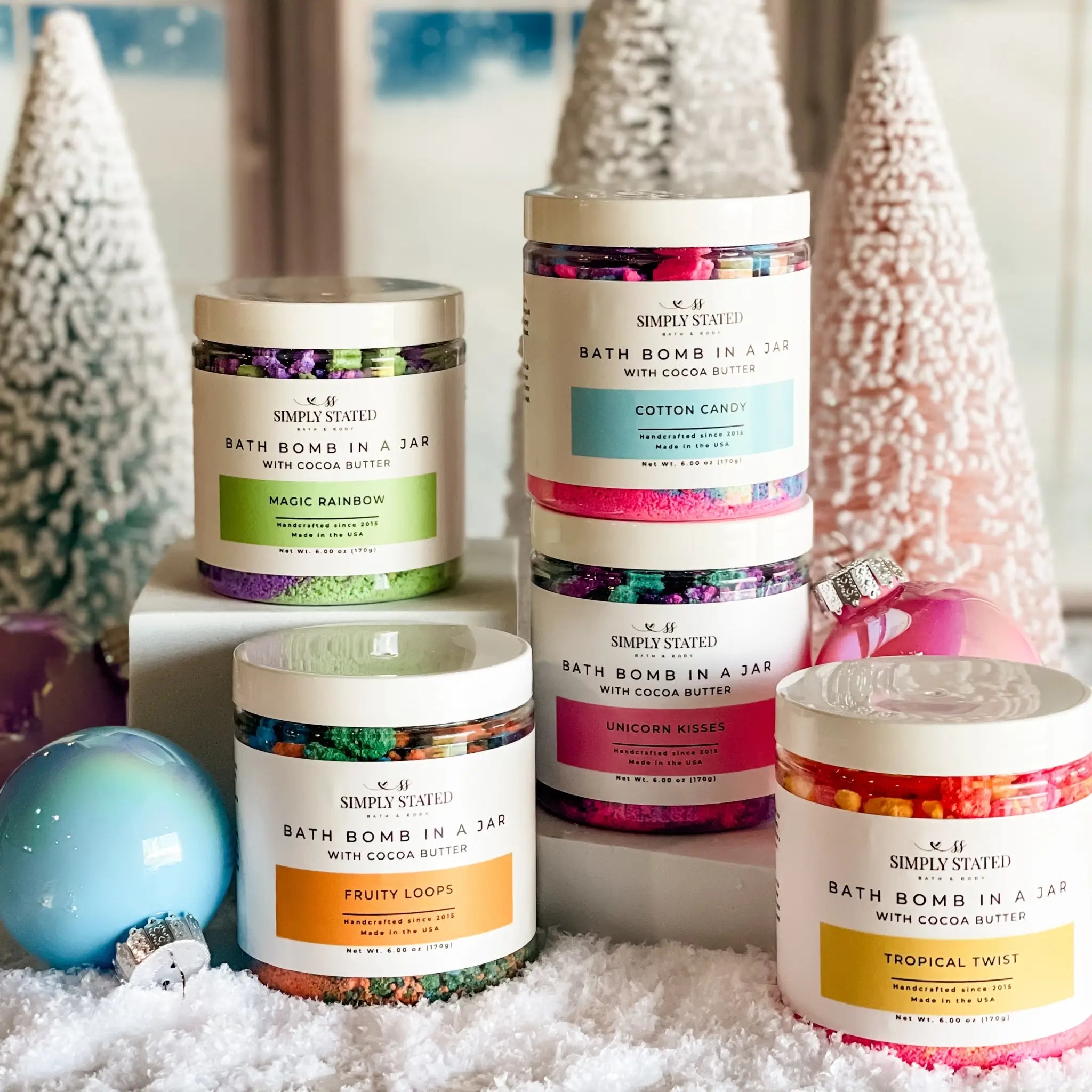 Bath bomb jars with colorful labels on a snowy surface with Christmas trees in the background