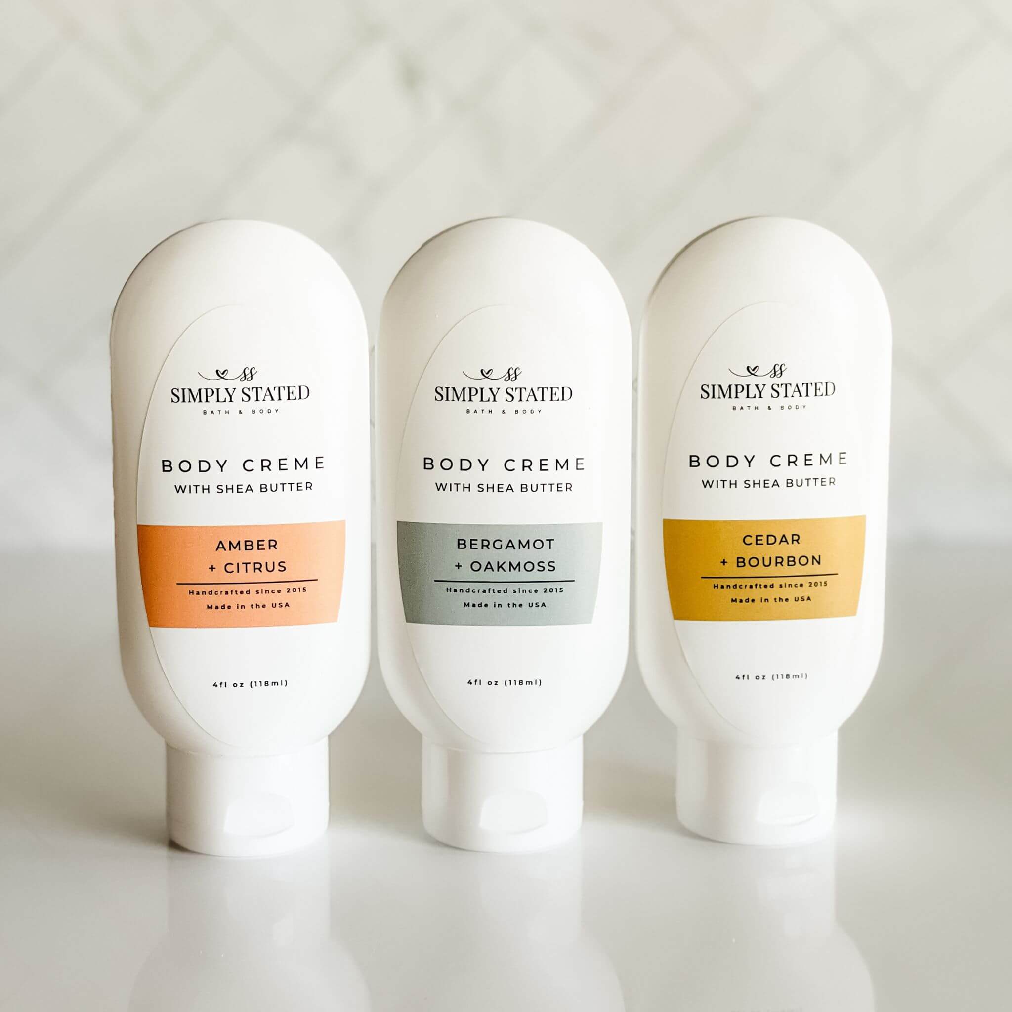 Group of 4 oz men's body creme bottles in three scents: Amber + Citrus, Bergamot + Oakmoss, and Cedar + Bourbon.