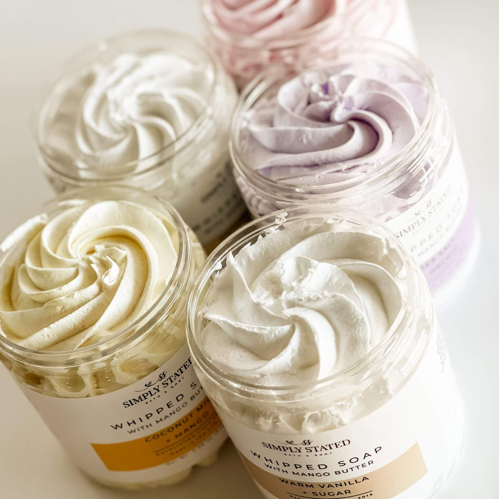 Open jars of Signature Line Whipped Soaps showcasing creamy whipped textures in vibrant colors, including Coconut Milk + Mango, Sweet Berry + Vanilla, Warm Vanilla + Sugar, Oatmeal Milk + Honey, and Sea Salt + Orchid.