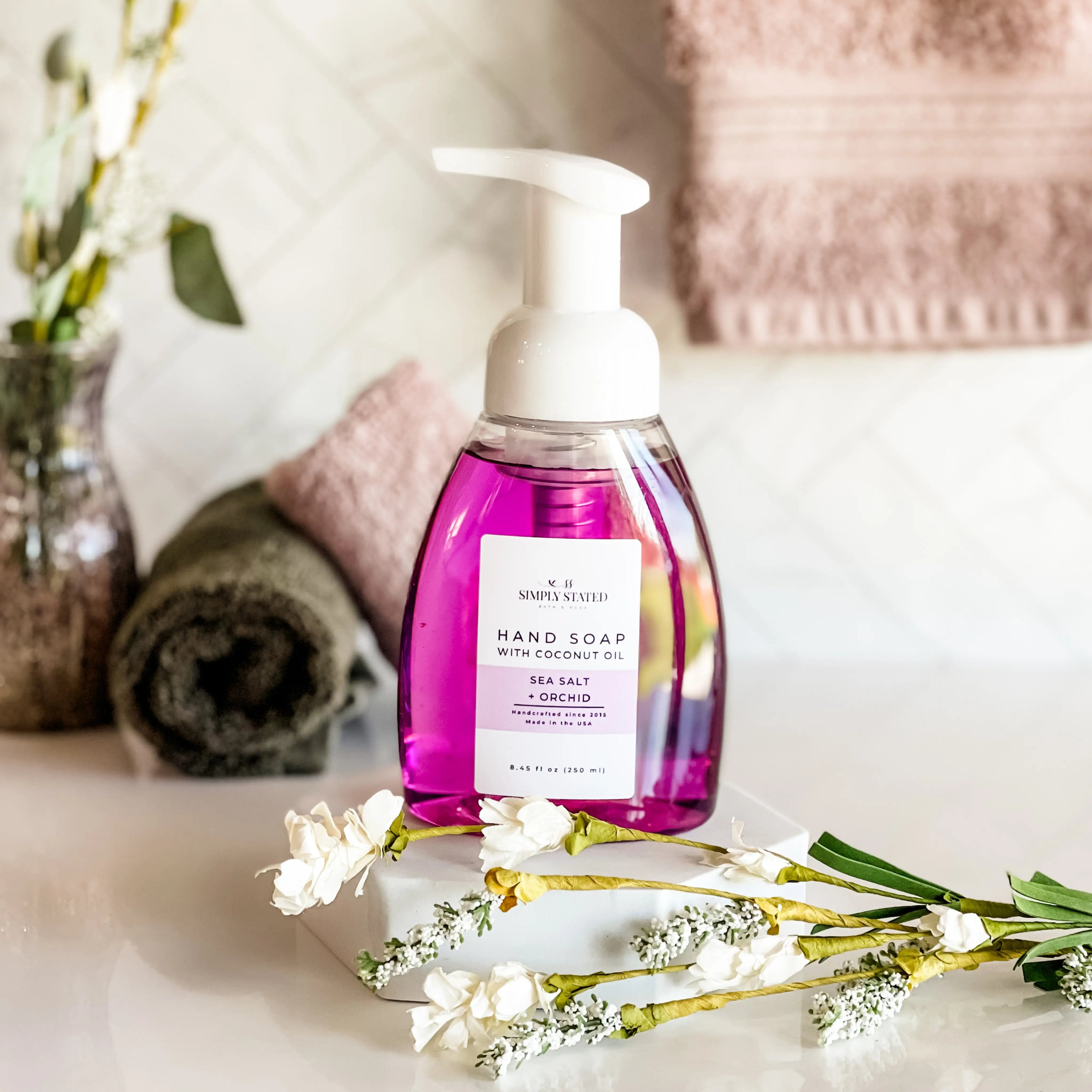 Purple hand soap bottle with pump on a marble surface with flowers and towels in the background