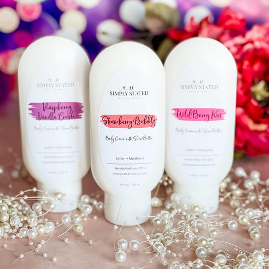 A trio of Sweet Temptations Body Cremes—Raspberry Vanilla Crush, Strawberry Bubbly, and Wild Berry Kiss—arranged on a blush-pink background with pearl accents, surrounded by soft floral decor.