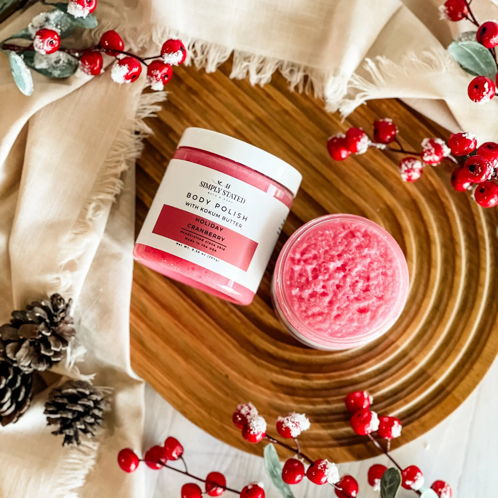Body polish jar with pink scrub on a wooden board surrounded by red berries and pine cones.