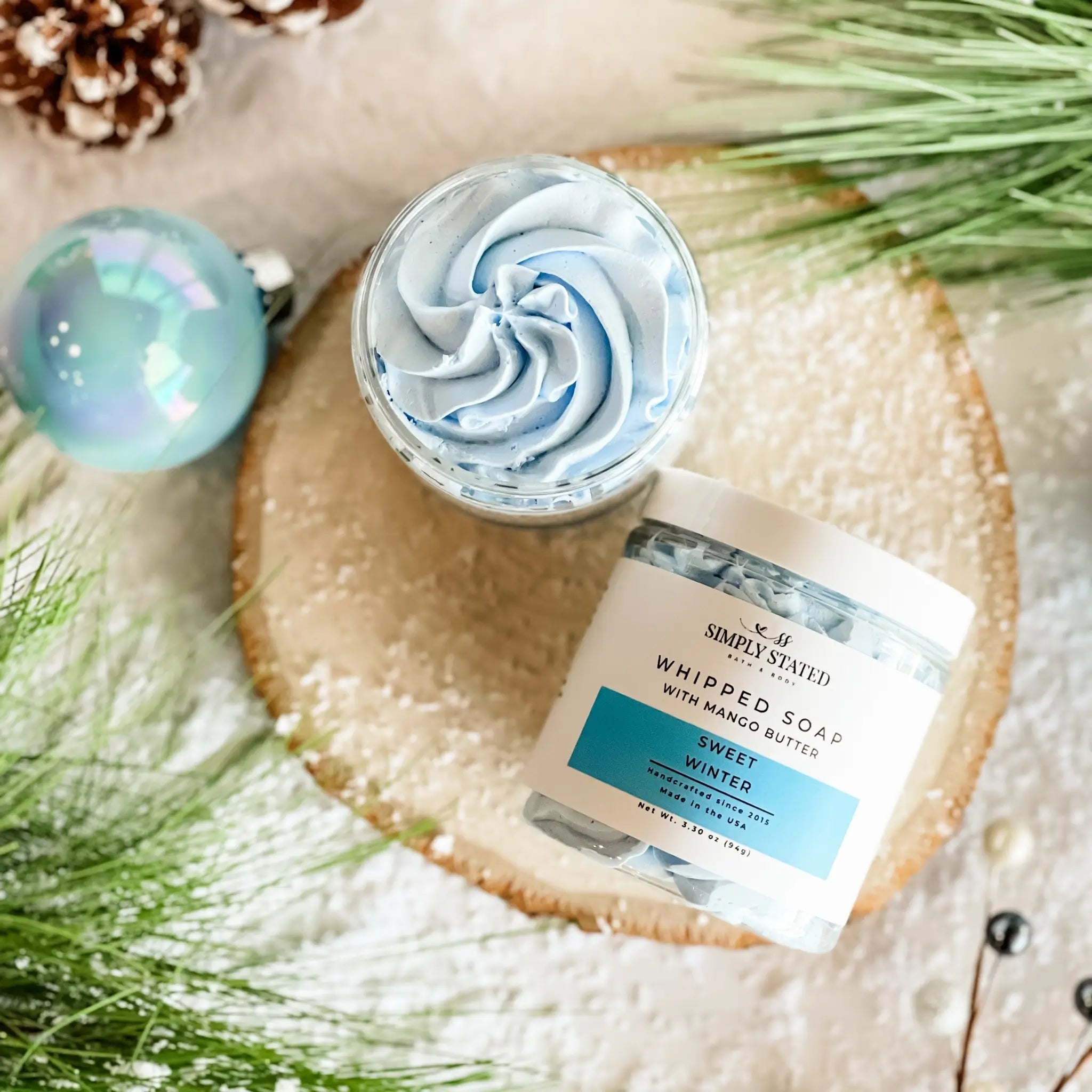 Whipped soap with a decorative label on a textured surface with greenery and a pine cone.