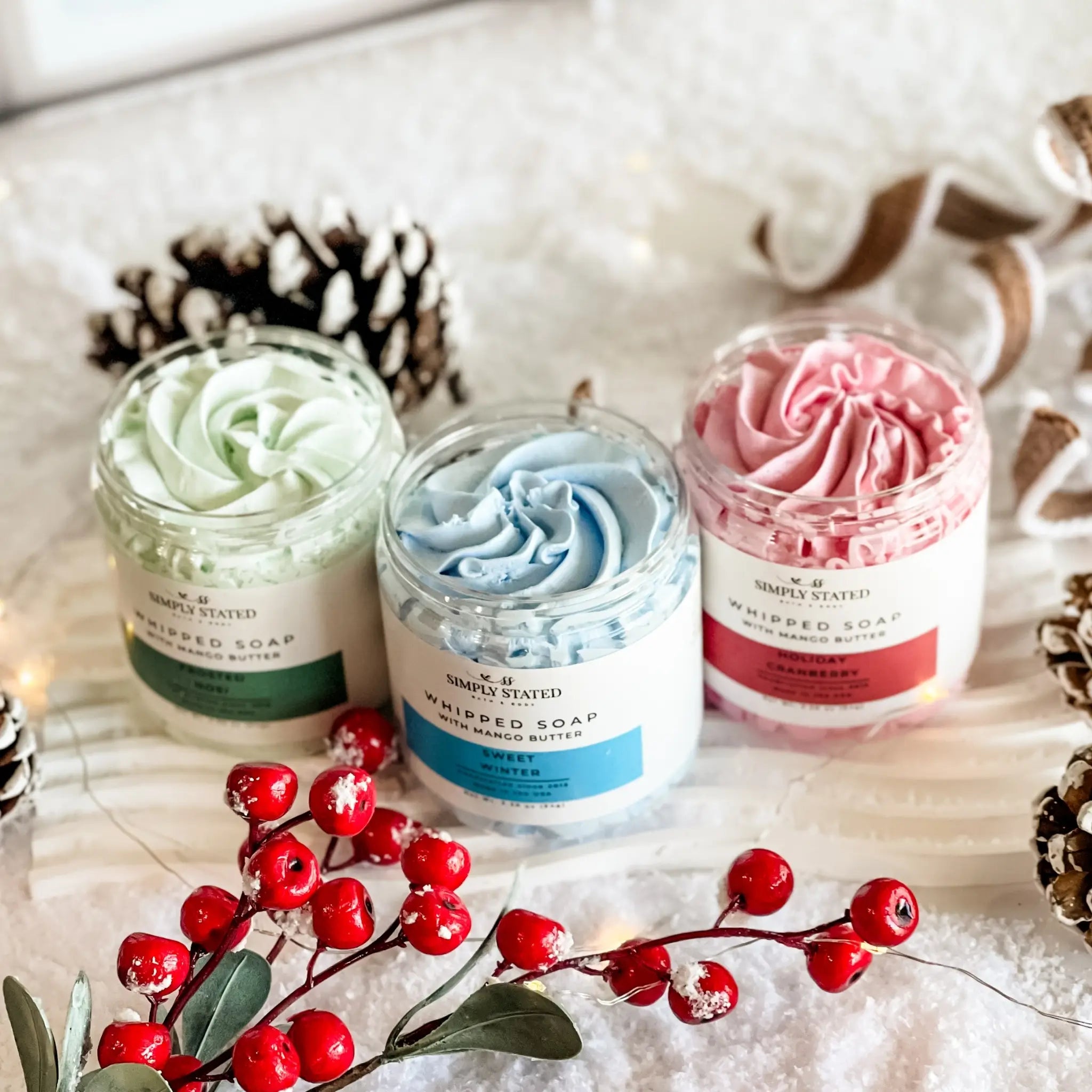 Three jars of whipped soap with decorative berries and pine cones on a textured surface.